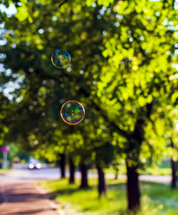 Two coloured translucent soap bubbles floating outdoors in the air on sunny evening with trees and car in the background.