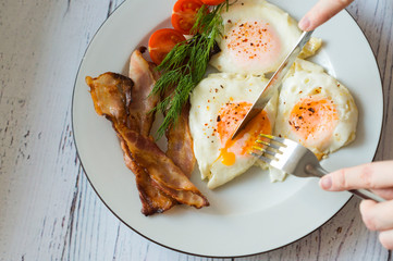 Fried eggs with bacon are cut with a knife and fork