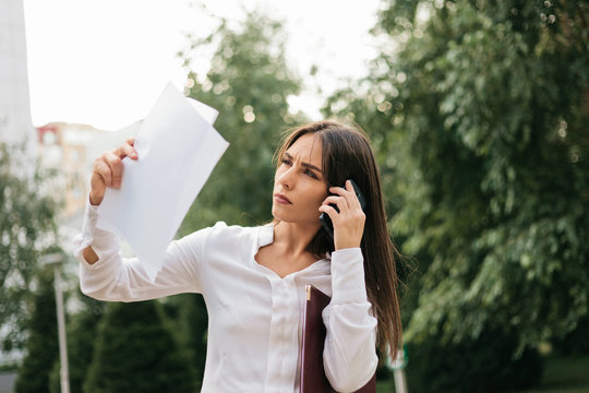 Young attractive female manager holds sheets of paper talking on the phone outdoors in the city. All in business