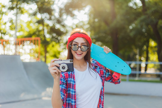Stylish hipster girl holding a skateboard and retro camera in hands on a bright sunny day. Youth concept. Summer lifestyle image of trendy pretty young girl