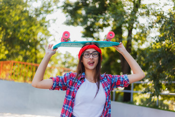 Funny young hipster woman dressed in stylish clothes holds skateboard in her hand in skatepark at...