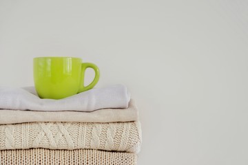 A green cup with a hot drink (coffee, tea, cocoa, cappuccino) on a stack of neatly folded knitted sweaters on a beige background. The concept of comfort, warmth, coziness, autumn-winter season.