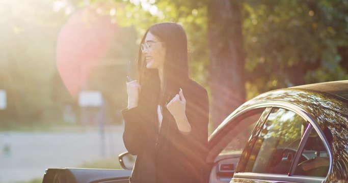 Successful caucasian brunette girl in jacket cheering up with cool news reading on smartphone standing by black car in the street.