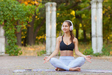 Fit woman in sports clothes sitting in lotus pose in the park and listening to music on headphones.  Healthy lifestyle. Yoga and relax concept. Look away