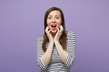 Amazed surprised young woman in casual striped clothes posing isolated on violet purple background in studio. People lifestyle concept. Mock up copy space. Keeping mouth open, putting hands on face.
