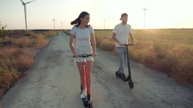 A guy and a young girl at sunset ride on electric scooters on the background of wind power generators. Alternative energy.