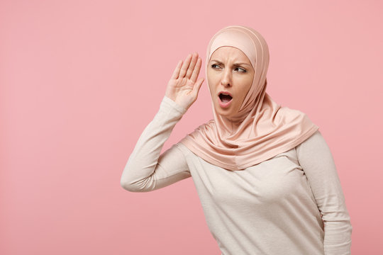 Curious young arabian muslim girl in hijab light clothes posing isolated on pink background in studio. People religious Islam lifestyle concept. Mock up copy space. Try to hear you with hand near ear.