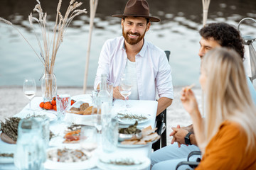 Group of young friends having fun during a festive dinner at the beautifully decorated table near the lake outdoors