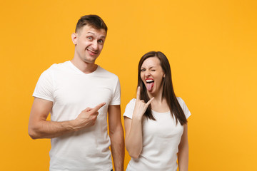 Young couple friends guy girl in white empty blank t-shirts posing isolated on yellow orange background. People lifestyle concept. Mock up copy space. Pointing finger, depicting heavy metal rock sign.
