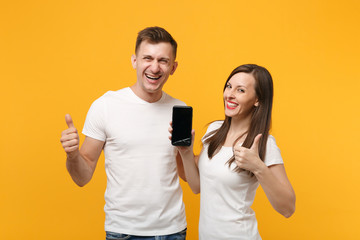 Funny couple friends guy girl in white t-shirts posing isolated on yellow orange background. People lifestyle concept. Mock up copy space. Hold mobile phone with blank empty screen, showing thumb up.