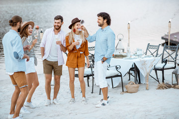 Group of young friends having fun, hanging out together with wine glasses during a festive party on the beach with dinning table on the background