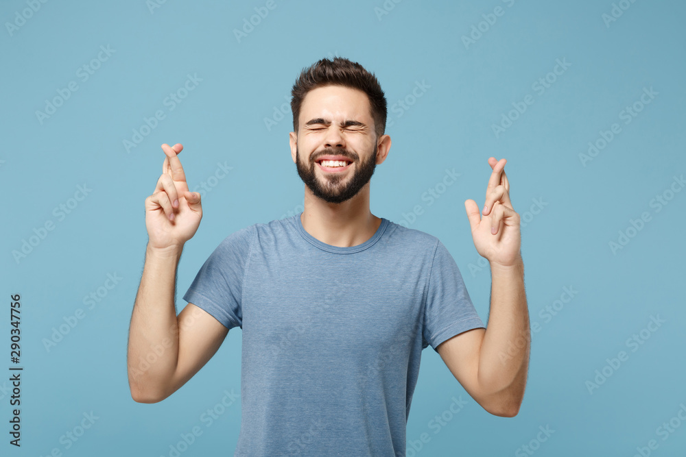 Wall mural Young funny man in casual clothes posing isolated on blue wall background. People lifestyle concept. Mock up copy space. Waiting for special moment, keeping fingers crossed, eyes closed, making wish.