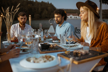 Friends having fun during a festive dinner at the beautifully decorated table with delicious meals on the beach at dusk