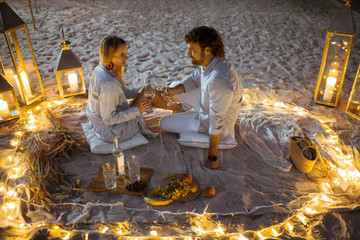 Couple having a romantic dinner, hugging and clinking wine glasses at the beautifully decorated place illuminated with different lights the sandy beach at dusk