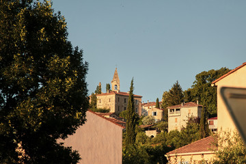 Fototapeta na wymiar View of the old town of Barjac, southern France. Green trees, roofs of buildings and stone church with a bell tower on a hill at sunset light