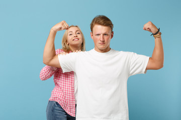 Young couple two friends guy girl in white pink empty blank design t-shirts posing isolated on pastel blue background in studio. People lifestyle concept. Mock up copy space. Showing biceps, muscles.