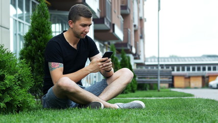 Attractive man use mobile phone while sitting on the grass near the hotel, app, chatting with friends