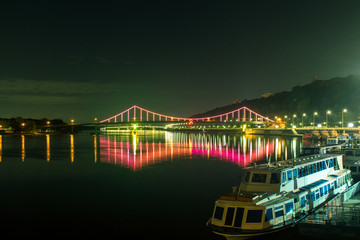 Fototapeta na wymiar A bridge glowing with colorful lights over a river. The bridge over the river and the ship standing on the pier at night