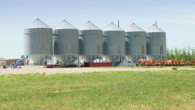 Wide shot of many large steel grain storage silos on Canadian farmland in the summer as a transport truck drives by in the background.