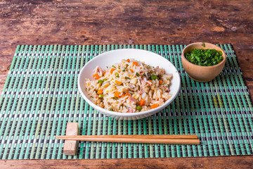 Cantonese rice is a traditional Chinese dish of fried rice, eggs, ham, peas, carrots and spices in a bowl on a bamboo napkin, chopsticks, top view