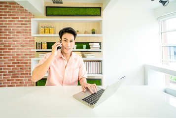 A Asian Handsome businessman 24s used mobile phone and working with laptop on desk in workplace near window,A handsome 24 year old Asian male student used phone and a laptop on the table in the house.