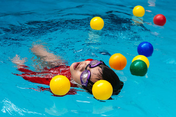 Young boy floating in the swimming pool with colored plastic balls around him.