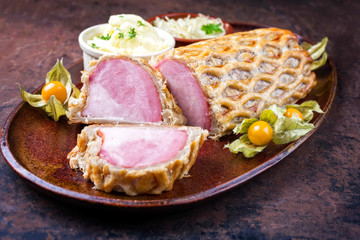 Gebackener Kasseler Schweinerücken im Blätterteig mit Sauerkraut und Kartoffelsalat als closeup angeboten auf einem Teller mit Physalis Früchten