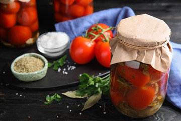 Pickled tomatoes in glass jars and products on black wooden table