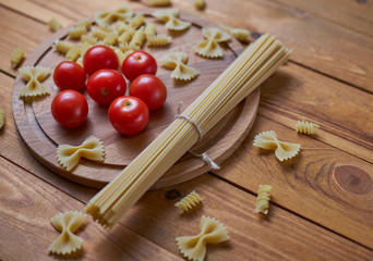Ripe and juicy cherry tomatoes with different types of raw pasta (spaghetti, farfalle, rotini) on a wood background.