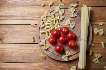 Top view image of different types of raw pasta (spaghetti, farfalle, fusilli) with cherry tomatoes on a wooden background.