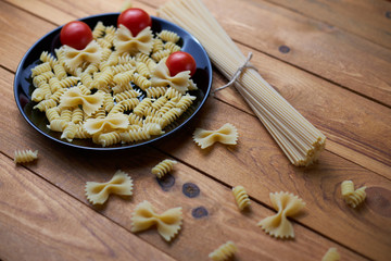 Cherry tomatoes with different types of raw pasta (spaghetti, farfalle, fusilli) on a wooden background.