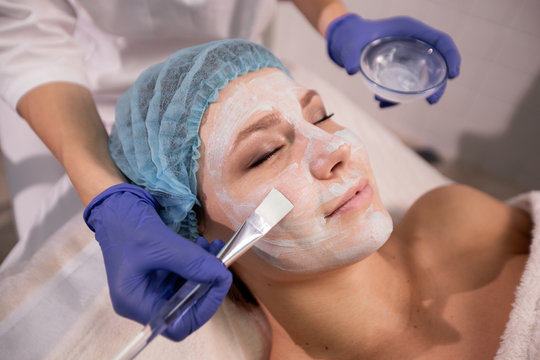 Close up of cosmetologist's hands in blue gloves applying a medical mask to the patient's face with a brush. Woman closed her eyes and enjoyed the process of rejuvenation