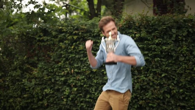 excited young casual man celebrates succeess while holding a trophy cup, he jumps and cheers