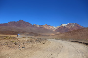 Valley with wide dusty road with light brown soil. Blue clear sky and colored mountains in the horizon. An off road vehicle running and raising dusty. Bolivia highlands desert.