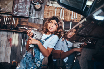 Fototapeta na wymiar Little cute girl is posing with pneumatic drill for photographer at auto service workshop.