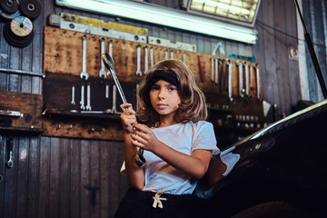 Fototapeta na wymiar Portrait of little cheerful girl with big wrench in hands near shiny gar at auto service.