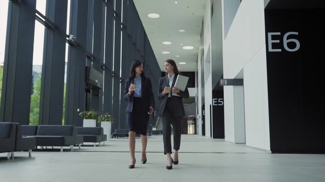 Full length shot of two businesswomen, young professional and her middle aged mentor, walking down office hall together and talking in slow motion