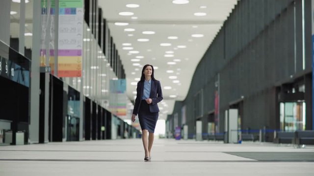 Lockdown wide shot of confident middle aged woman in suit and high heels shoes walking down office hall or airport lobby in slow motion