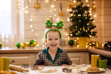 Kids bake Christmas gingerbread. Happy beautiful girl in pajamas preparing dough for new year cookies to celebrate Christmas. White kitchen with a Christmas tree and lights on a snowy winter day.