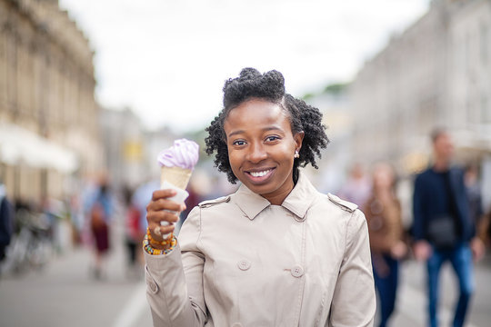 Lifestyle street girl. Afro-American girl in a light brown coat stands on a city street. Tourist walk of a stylish woman. She is eating pink strawberry ice cream.
