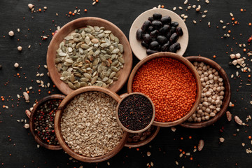 top view of wooden bowls and plates with raw lentil, oatmeal, beans, chickpea, quinoa, pumpkin seeds and peppercorns on dark surface with scattered grains