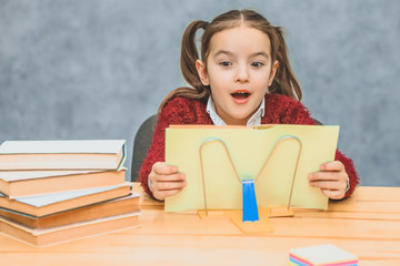 Child and school books on the background of a gray wall on the table. Performs a homework while reading a book. A schoolgirl sits on her desk with textbooks.