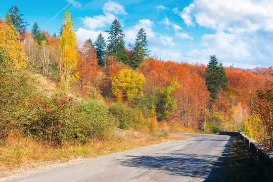 country road through forest in mountains. beautiful transportation autumn scenery in the morning. trees in colorful foliage. concrete border along the old cracked asphalt surface. clouds on the blue s