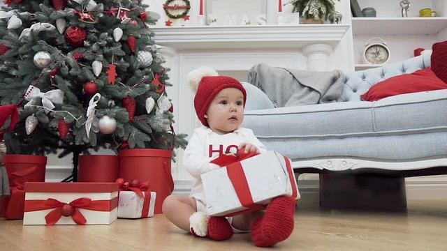 Christmas picture of a sweet little baby in christmas clothes sitting on the parquet and holding a gift near the christmas tree