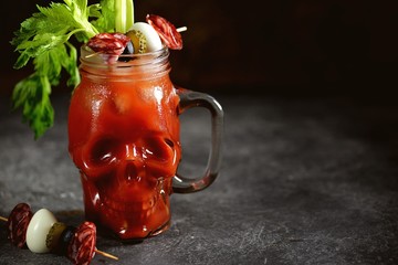 Bloody Mary cocktail in glass skull with celery sticks, pink salt, lime and canapes from canned vegetables. Halloween drink. 