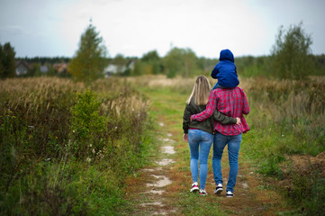 A young family in bright casual clothes walks away along a country road in the field. Little son on father's shoulders. Back view