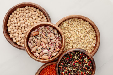 top view of wooden bowls with oatmeal, red lentil, beans, peppercorns and chickpea on white marble surface