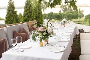 Wedding table setting decorated with fresh flowers in a brass vase. Wedding floristry. Banquet table for guests outdoors with a view of green nature. Bouquet with roses, eustoma and eucalyptus leaves