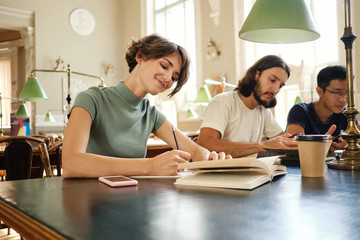 Young attractive female student with book happily working on study project in library of university