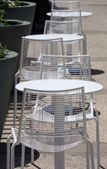 A row of metal outdoors white chairs and tables in set along large flower planters along a street curb